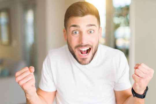 Young handsome man wearing casual white t-shirt at home celebrating surprised and amazed for success with arms raised and open eyes. Winner concept.