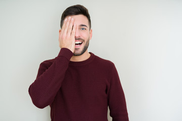 Young handsome man wearing a sweater over isolated background covering one eye with hand with confident smile on face and surprise emotion.