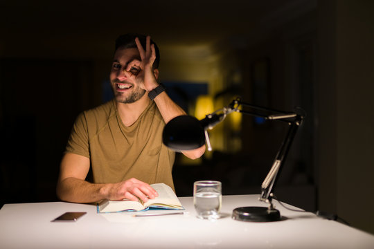 Young handsome student man studying at night at home with happy face smiling doing ok sign with hand on eye looking through fingers