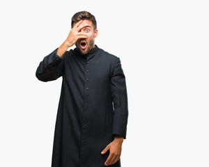Young catholic christian priest man over isolated background peeking in shock covering face and...