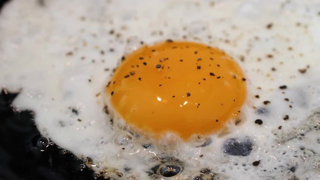 Close-up of egg frying in cast iron apn with black pepper. Pan and half speed slow motion.