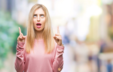 Young beautiful blonde woman wearing pink winter sweater over isolated background amazed and surprised looking up and pointing with fingers and raised arms.