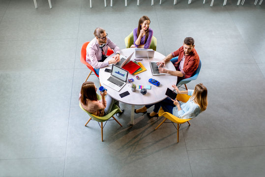 Team Of A Young Coworkers Dressed Casually Working Together With Laptops Sitting At The Round Table In The Office, View From Above