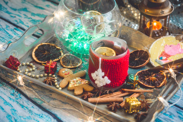New Year's still life with mug of tea with knitted cup cover, cookies, chocolate, dried fruit and seasonings with lights and bokeh. Photo toned and with vignette