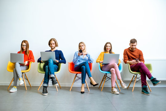 Young coworkers working with gadgets sitting in a row on the colorful chairs on the white wall background indoors