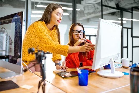 Two young creative women dressed in sweaters working with computer in the modern office
