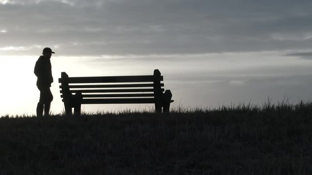 Silhouetted person walks up to empty park bench to sit and enjoy the scenery.