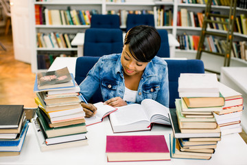 Smart african american young woman dressed in casual wear preparing for studying seminar in library.Intelligent dark skinned hipster student writing and reading