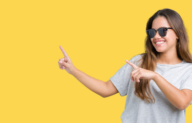 Young beautiful brunette woman wearing sunglasses over isolated background smiling and looking at the camera pointing with two hands and fingers to the side.