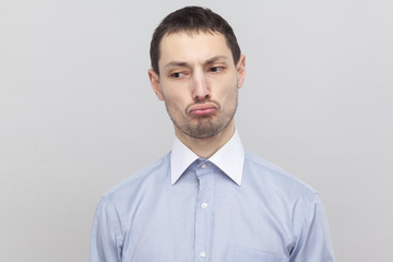 Closeup portrait of sad dissatisfied handsome bristle businessman in classic light blue shirt standing and looking away with pout lips. indoor studio shot, isolated on grey background copyspace.