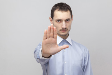 Portrait of serious handsome bristle businessman in classic light blue shirt standing and showing stop gesture sign. indoor studio shot, isolated on grey background copyspace.