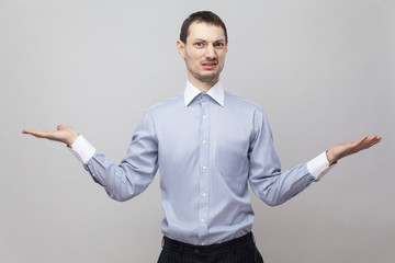 I don't know, which one to choose. Portrait of confused bristle businessman in blue shirt standing with raised arms and looking at camera. indoor studio shot, isolated on grey background copyspace.