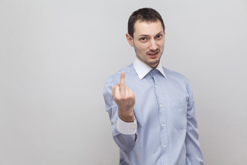 Portrait of angry handsome bristle businessman in classic blue shirt standing, looking at camera with middle finger fuck sign with poker face. indoor studio shot, isolated on grey background copyspace