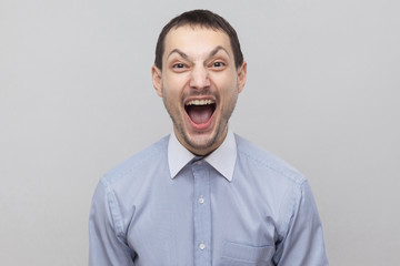Portrait of handsome bristle businessman in classic light blue shirt standing with open mouth, screaming and looking at camera. indoor studio shot, isolated on grey background copyspace.