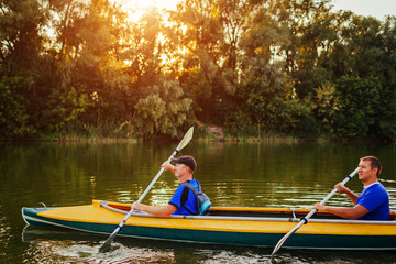 Young men rowing kayak on river at sunset. Couple of friends having fun canoeing in summer