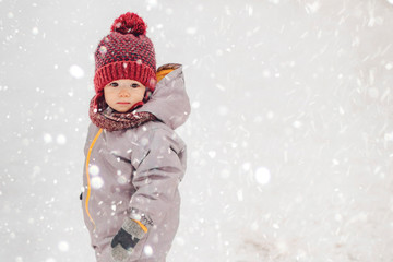 Portrait of a cute baby dressed in a gray jacket and a red hat that walks through the snow covered park  enjoying first snow blowing