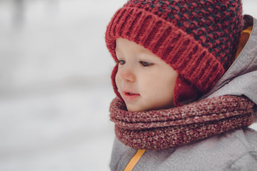 Portrait of a cute baby dressed in a gray jacket and a red hat that walks through the snow covered snow park. She smiles one in the photo during the snowfall