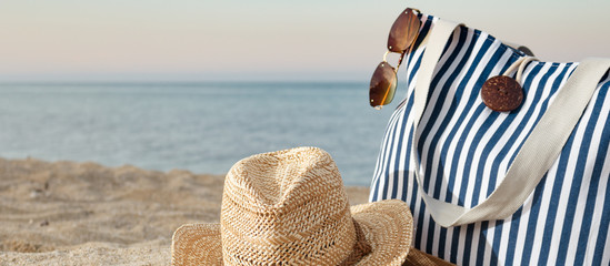 Striped summer bag with straw hat and sunglasses closeup on beach and calm sea background. Sunset...