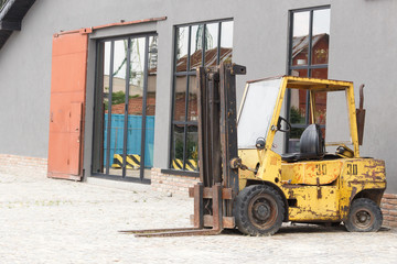 Old yellow, black, rusty scratched forklift truck parked on road near big industrial warehouse converted into modern restaurant and sidewalk in Varna, Bulgaria. Vehicle also named fork truck.