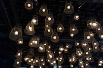 Many vintage incandescent glowing light bulbs with net lampshade hanging from the ceiling on the dark background texture, restaurant interior