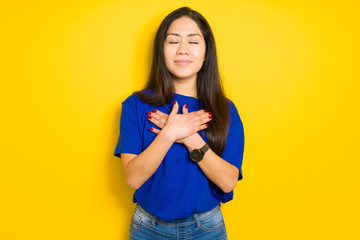 Beautiful brunette woman wearing blue t-shirt over yellow isolated background smiling with hands on chest with closed eyes and grateful gesture on face. Health concept.