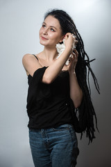 Fototapeta premium Cute and funny dreaded hipster girl wearing a black top and blue jeans and big snowflake earrings posing on a staircase against a white background.