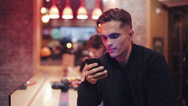 Young Attractive Man with Beautiful Smile is Using Smartphone at Evening. He sitting in a bar or restaurant near neon signage. Close up