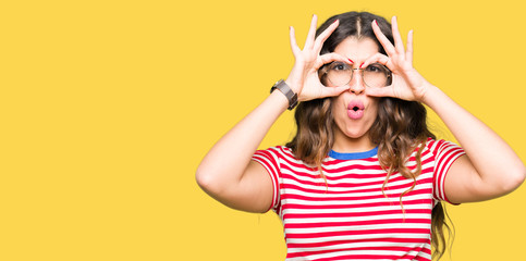 Young beautiful woman wearing glasses doing ok gesture like binoculars sticking tongue out, eyes looking through fingers. Crazy expression.