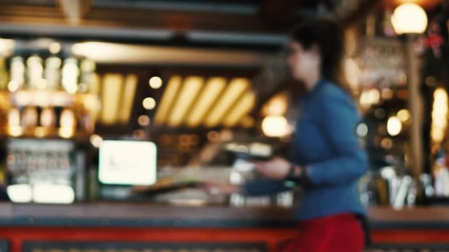 Blurred image of the interior of a large beautiful restaurant with bright lighting. The waitress comes to the working bar and the bartender, gives visitors a menu and goes for a drink. Defocused staff