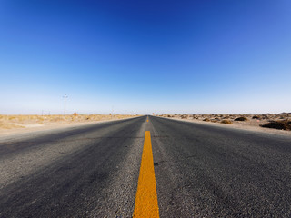 Fototapeta na wymiar Centered view of an endless asphalt road in desert, low angle straight rough road with blue sky background, magic world.
