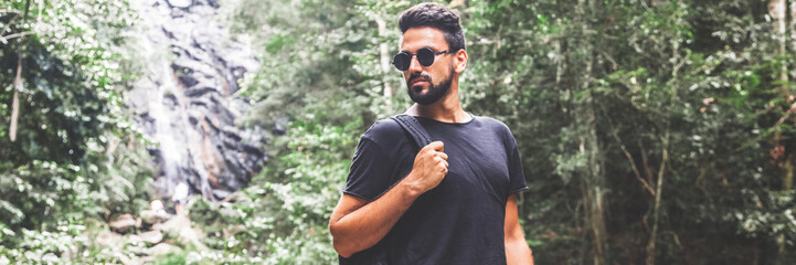 Handsome young stylish man in black t-shirt and sunglasses is engaged in trekking in the green jungle.