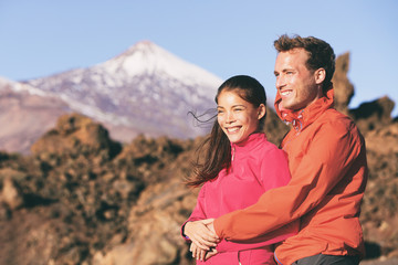 Happy hiking couple relaxing in mountain nature hugging. Interracial lovers hikers smiling, Asian woman, Caucasian man wearing winter jackets for cold spring. Outdoor living lifestyle.
