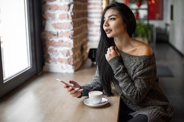 Beautiful girl uses a phone and drinks coffee, sitting in a cozy cafe.