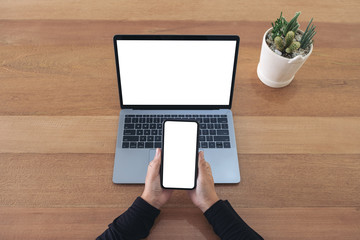 Top view mockup image of hands holding a blank white screen mobile phone and laptop on wooden table...