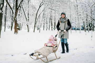 Portrait happy young mother with daughter, stand  with baby and a children sled outdoors on the background winter. Close up. Happy family walking in the park. Infant.