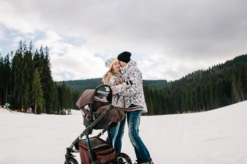 Young family couple hugging, stand near baby stroller on snow in Carpathian mountains. On background of forest and ski slopes. Close up. Winter nature. Hoist in  mountains. A man kisses a woman.