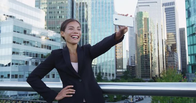 Smiling young businesswoman taking selfie with mobile phone against office buildings. Beautiful female professional is using smart phone in financial district. She is wearing suit in city.