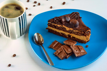 A piece of chocolate cake, and pieces of chocolate on a blue plate, next to a cup of coffee, on a light wooden background.