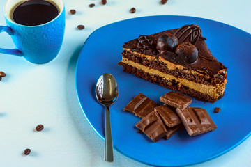 A piece of chocolate cake, and pieces of chocolate on a blue plate, next to a cup of coffee, on a light wooden background.