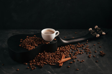 A cup of coffee is on a black ululele with scattered coffee beans, anise and cinnamon