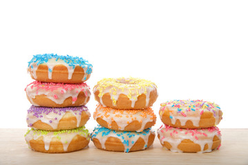 Many colorful frosted cake donuts with candy sprinkles stacked in rows laying on wood table...