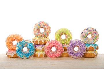 Many colorful frosted cake donuts with candy sprinkles stacked and up on side laying on wood table isolated on white background. Fun festive party food.