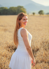 Fototapeta na wymiar Young woman in white dress looking back over her shoulder, smiling, afternoon sun lit wheat field behind her.