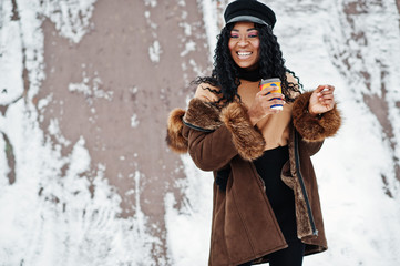 Fototapeta na wymiar African american woman in sheepskin coat and cap posed at winter day against snowy background with cup of coffee.