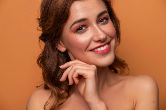 Cute caucasian brunette woman with curly hairstyle on orange background. Fresh clean skin with evening make up and freckles. Hands near her face. Close up portrait