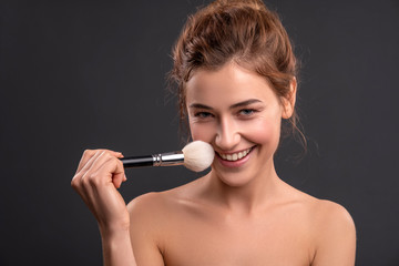 Obraz premium Beautiful brunette caucasian young woman prepare herself, applying powder on her cheeks with a brush. Clean, fresh, natural, flawless skin. Soft smile on her face. Close up on a neutral background