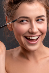 Beautiful brunette caucasian young woman prepare herself, applying mascara on her eyes with a brush. Clean, fresh, natural, flawless skin. Soft smile on her face. Close up on a neutral background