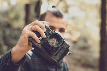 Hipster Travel Lifestyle concept: stylish bearded man, dressed in a shirt and with a hairdo Top Knot with retro photo camera in hands takes pictures in the woods