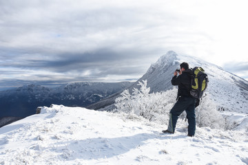 Fototapeta na wymiar Hiker with backpack and camera taking picture of snowy mountain. Winter mountain landscape, Happy tourist hiking in winter 