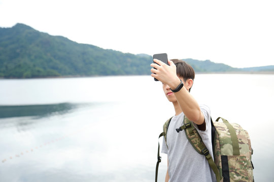 Asian traveler with backpack holding the smartphones to take photo themselves (Selfie) on a bridge by the lake. - image
